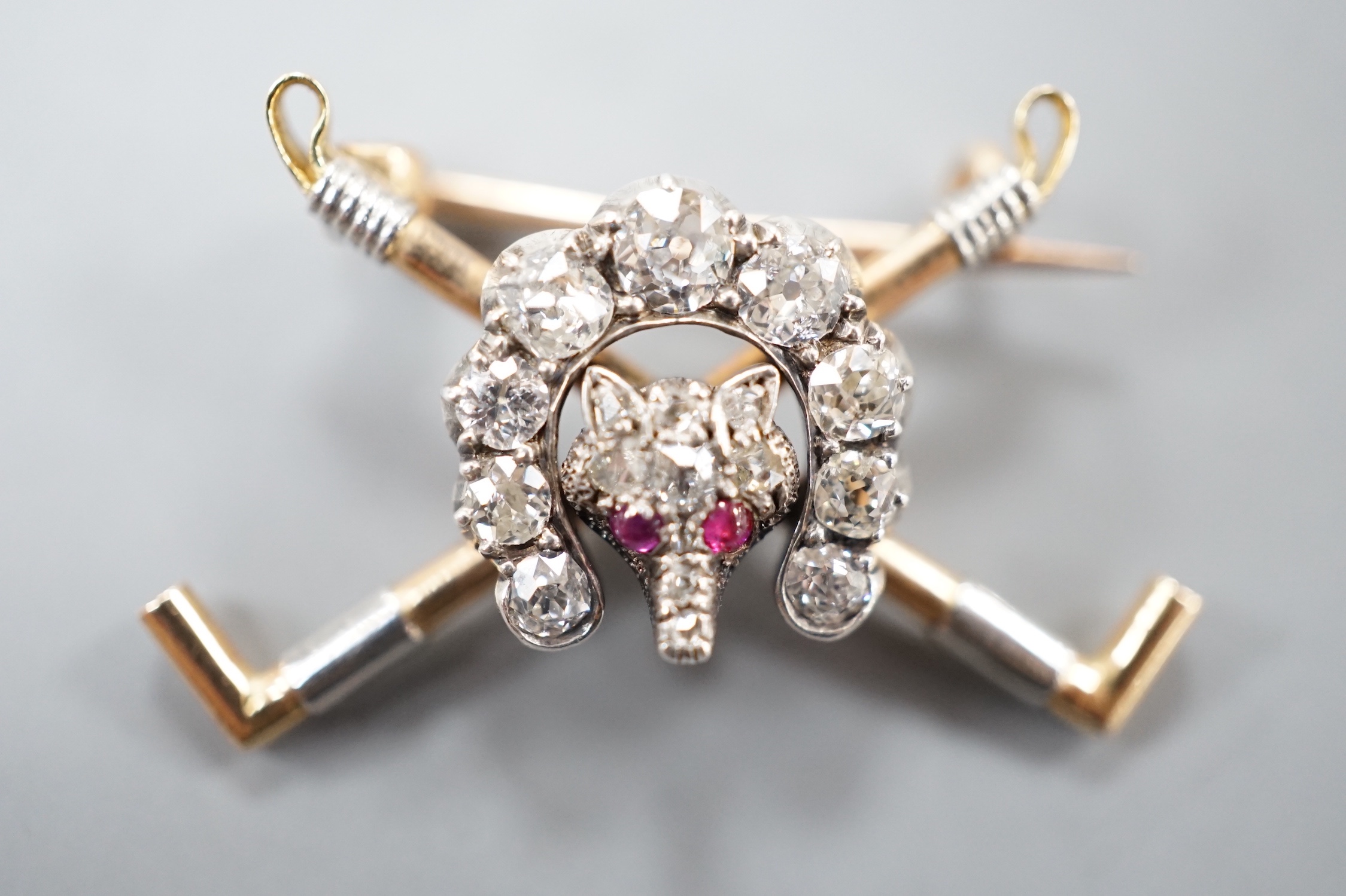 An Edwardian yellow metal, ruby and diamond set hunting brooch, modelled as a horseshoe and fox head upon crossed riding crops, 29mm, gross 4.2 grams.